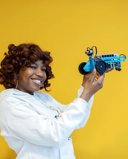 Student in a white hoodie smiling while holding a small blue robotic vehicle against a bright yellow background. Student in a white hoodie smiling while holding a small blue robotic vehicle against a bright yellow background.